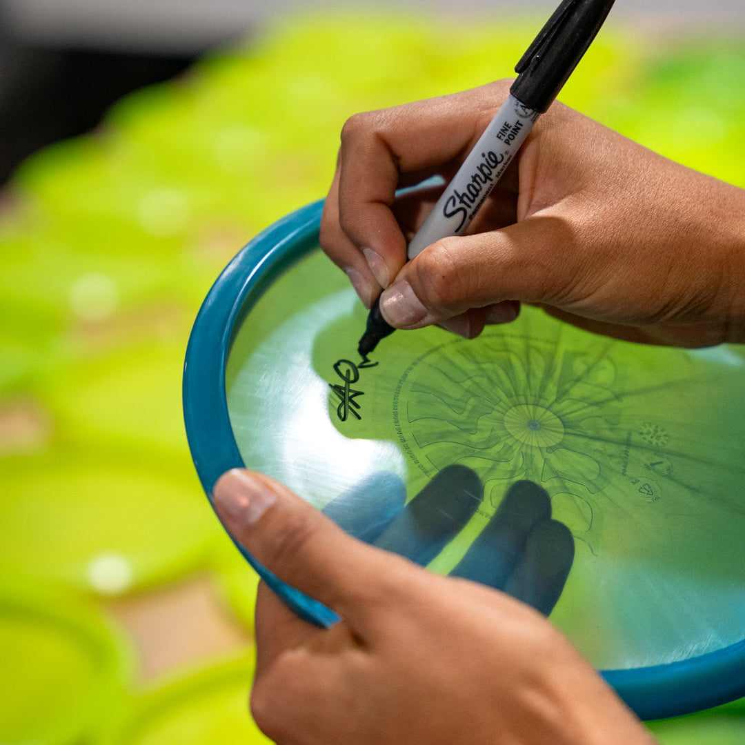 Maria Oliva signing her blue Frozen signature series trash panda midrange disc golf midrange